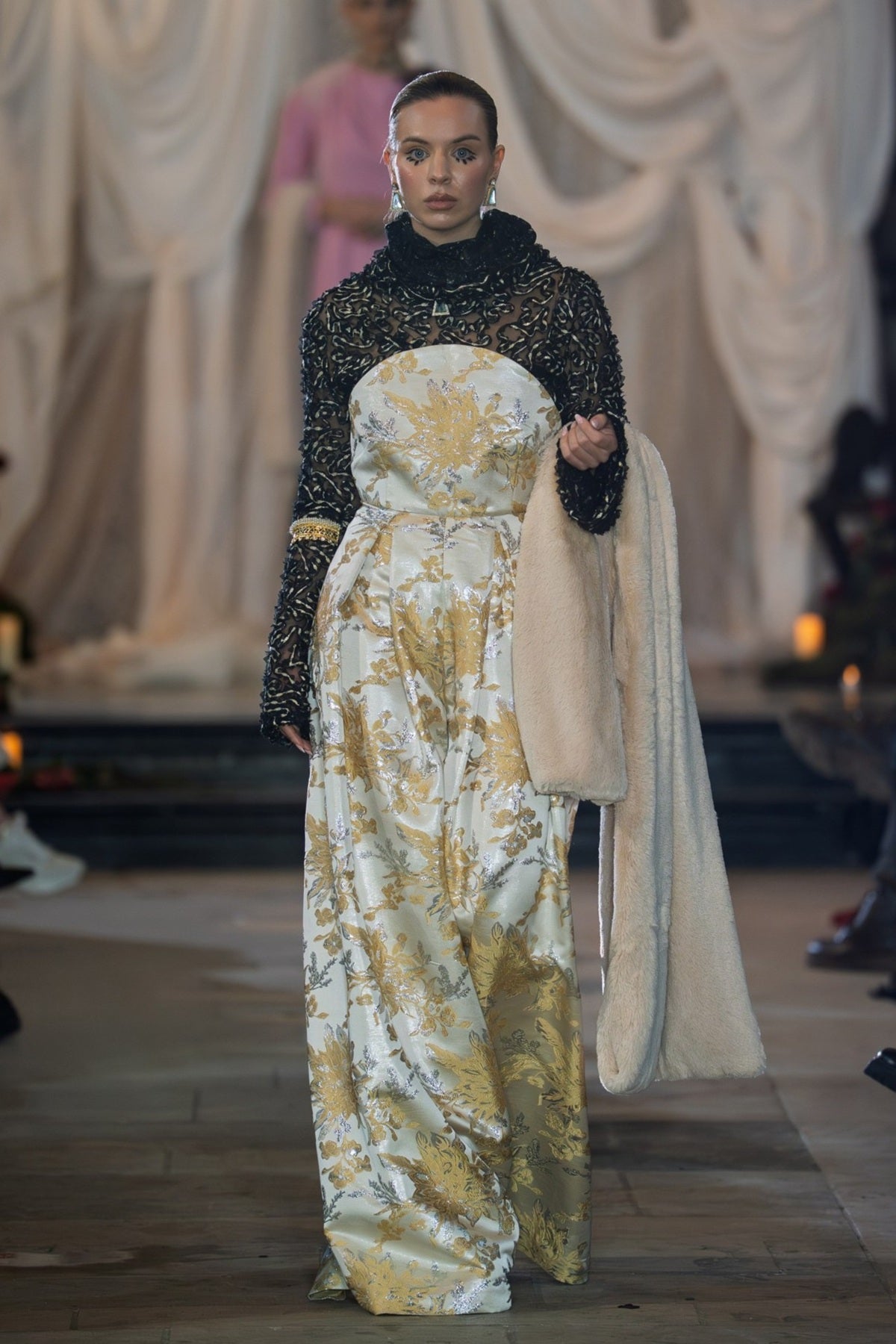 Model walking on a runway wearing a gold and white dress with a black top.