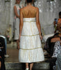 A model from behind wearing a white textured midi dress with gold trim and thin straps, walking down a fashion runway with an audience seated on both sides.