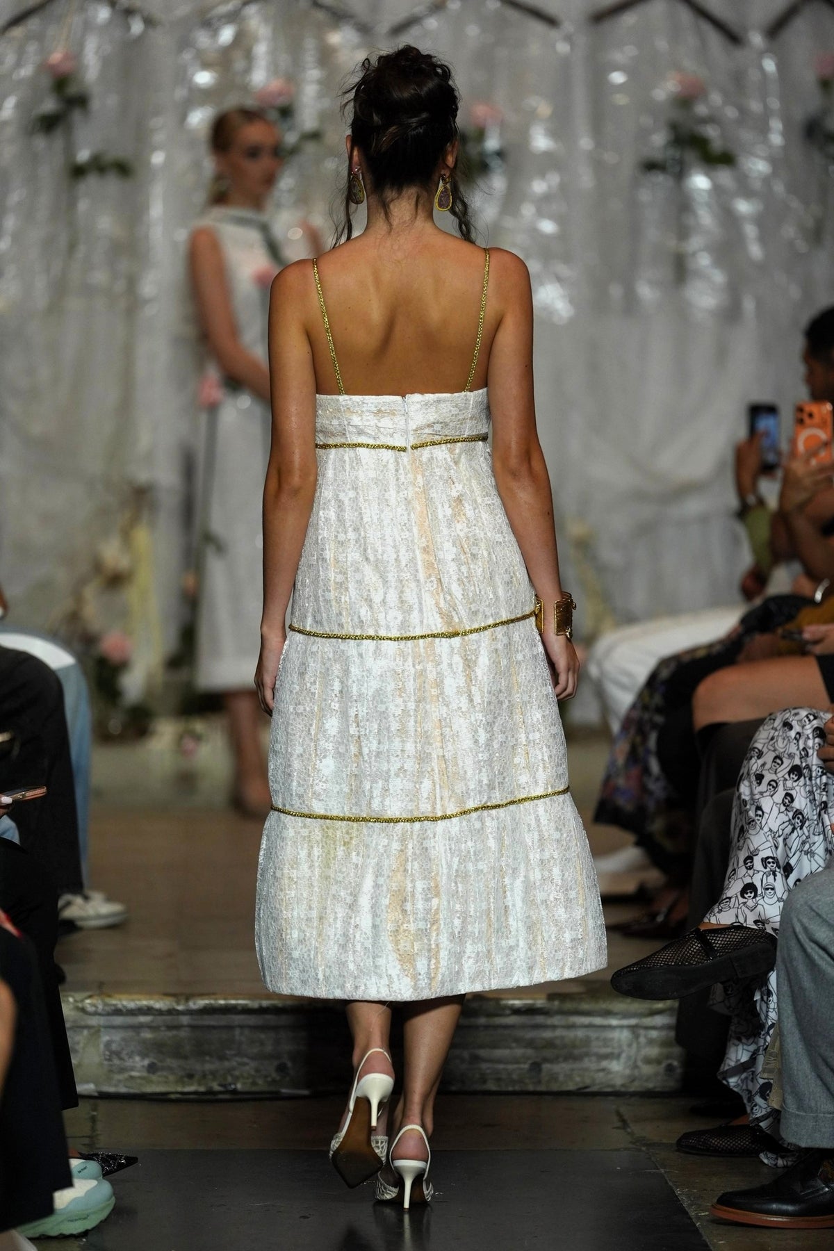 A model from behind wearing a white textured midi dress with gold trim and thin straps, walking down a fashion runway with an audience seated on both sides.