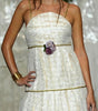 A model walks the runway wearing a strapless white textured midi dress with gold trim and a floral waist detail, set against a backdrop of hanging white garments.