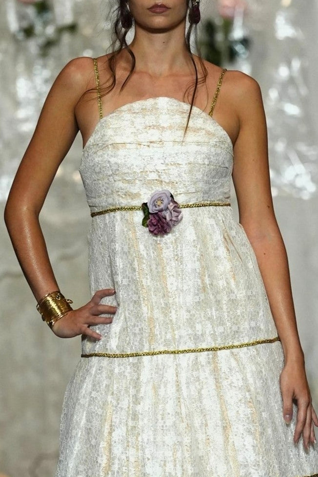 A model walks the runway wearing a strapless white textured midi dress with gold trim and a floral waist detail, set against a backdrop of hanging white garments.