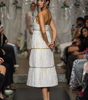 A model on a fashion runway wearing a white sleeveless midi dress with gold trim and a tiered skirt, photographed in a runway show with seated spectators on both sides.