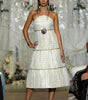 A model walks the runway wearing a strapless white textured midi dress with gold trim and a floral waist detail, set against a backdrop of hanging white garments.