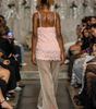 A model walks down a fashion runway from behind, wearing a light pink textured spaghetti-strap top and sheer beige wide-leg trousers with red high heels.