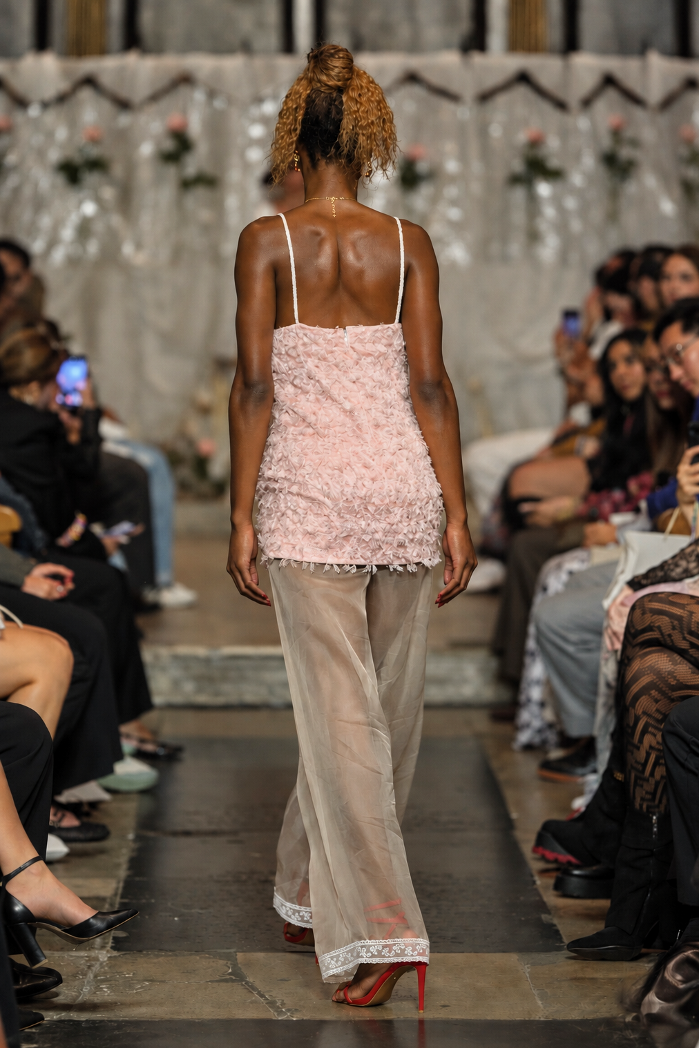 A model walks down a fashion runway from behind, wearing a light pink textured spaghetti-strap top and sheer beige wide-leg trousers with red high heels.