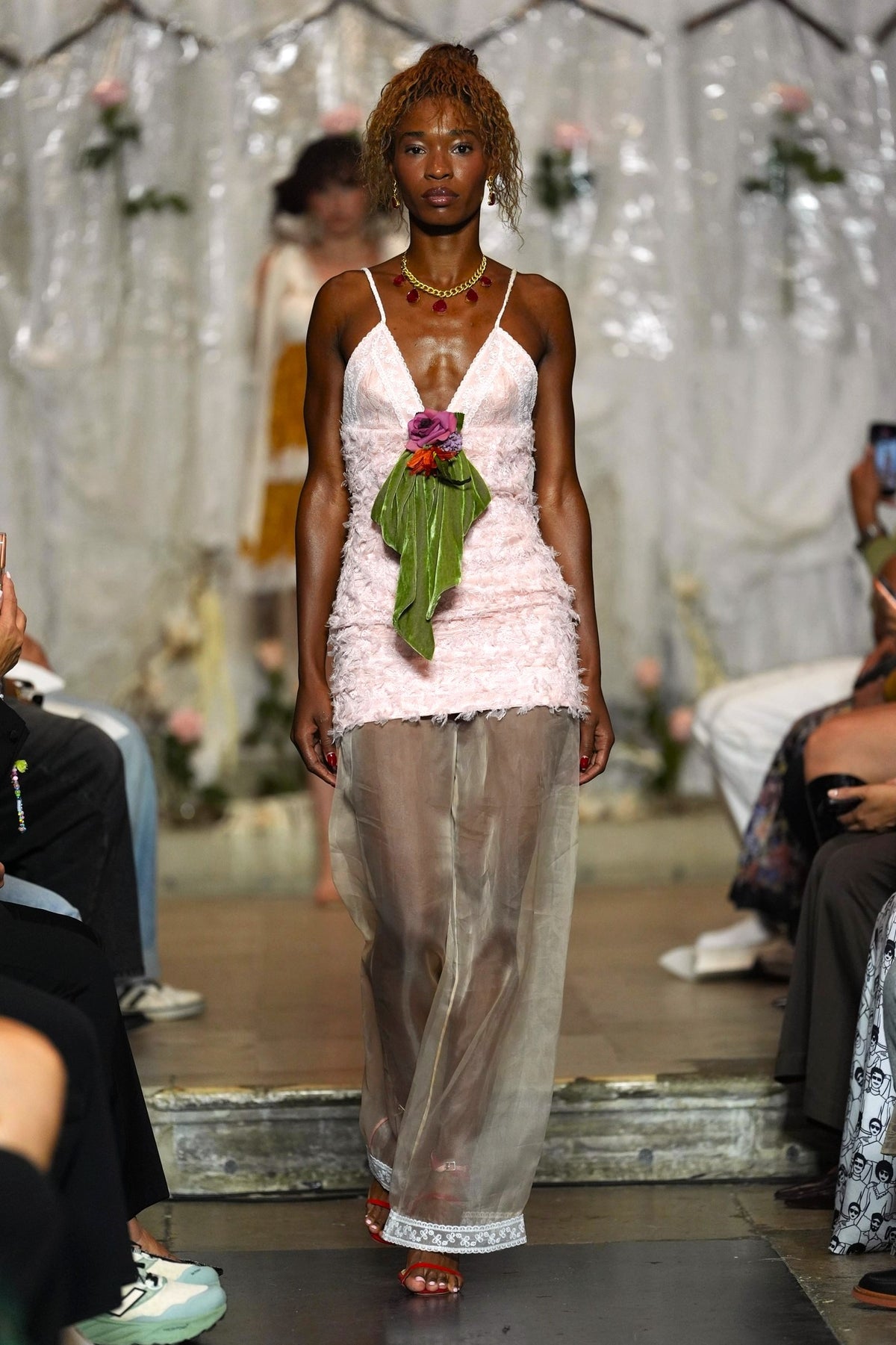 A model walks the runway wearing a light pink ruffled spaghetti-strap dress with sheer wide-leg tulle pants and a green leaf accent at the waist.