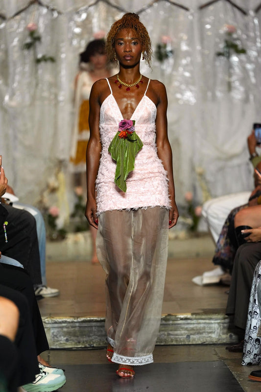 A model walks the runway wearing a light pink ruffled spaghetti-strap dress with sheer wide-leg tulle pants and a green leaf accent at the waist.