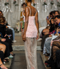 A model on a fashion runway viewed from behind, wearing a light pink textured mini dress with thin straps and a sheer, floor-length overlay skirt in a gray-beige tone, paired with red high heels.