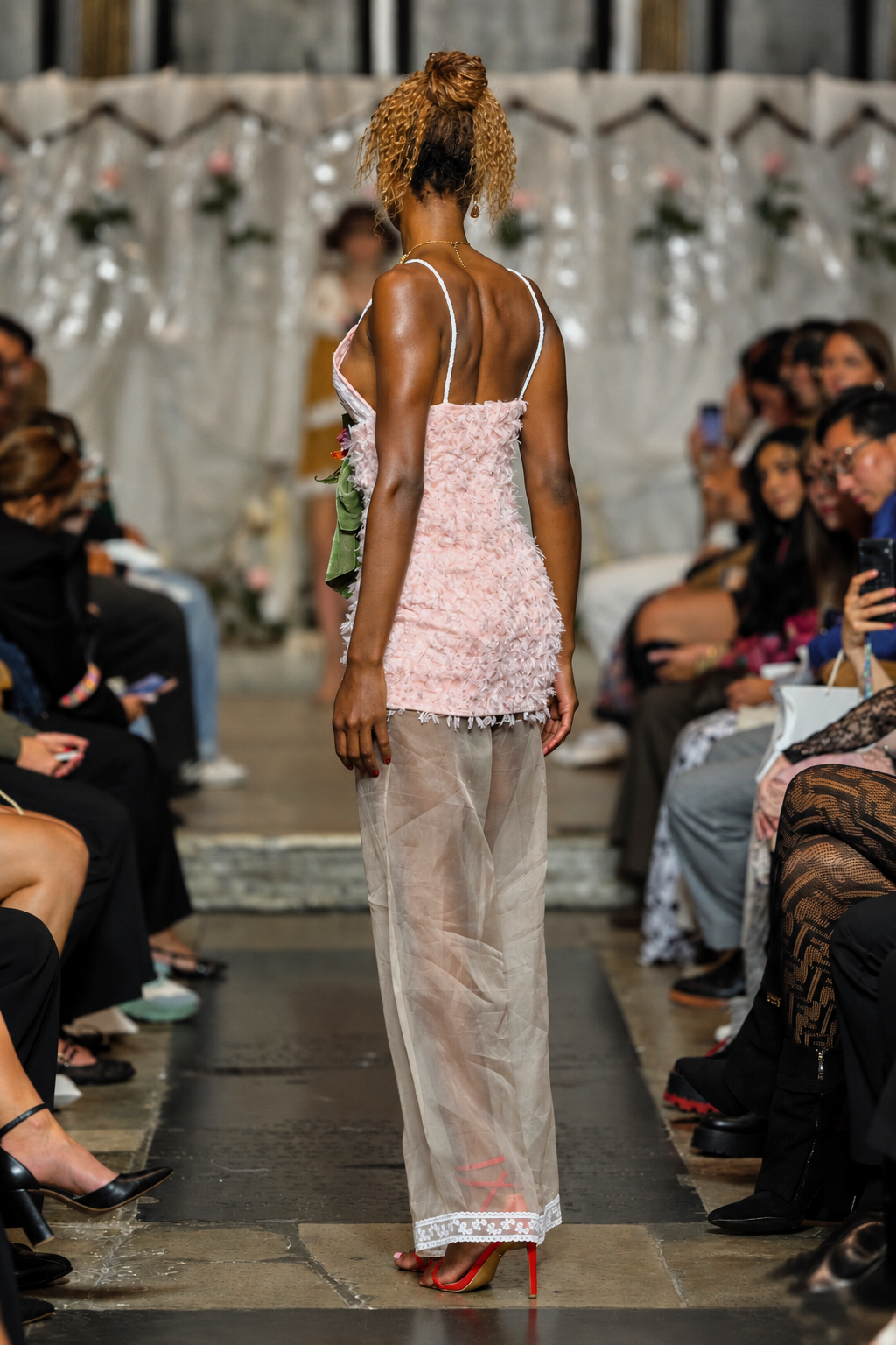 A model on a fashion runway viewed from behind, wearing a light pink textured mini dress with thin straps and a sheer, floor-length overlay skirt in a gray-beige tone, paired with red high heels.