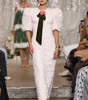 A model walks the runway wearing a light pink textured feather dress with puffed short sleeves, a dark green satin bow at the chest, and a pink rose accent.