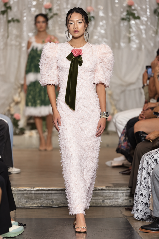 A model walks the runway wearing a light pink textured feather dress with puffed short sleeves, a dark green satin bow at the chest, and a pink rose accent.