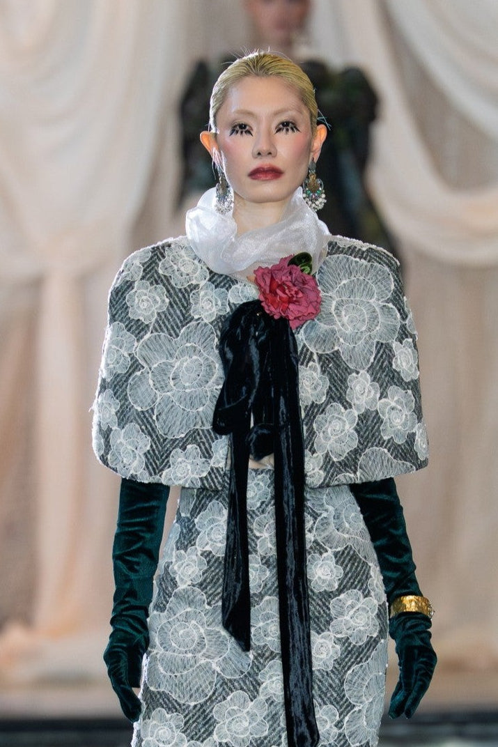 Model walking on a fashion runway wearing a patterned outfit with a floral accessory.