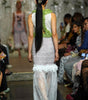 A model viewed from behind walking down a runway wearing a sleeveless white lace dress with a textured lime green bodice and a sheer white tulle overskirt. The runway is lined with spectators.