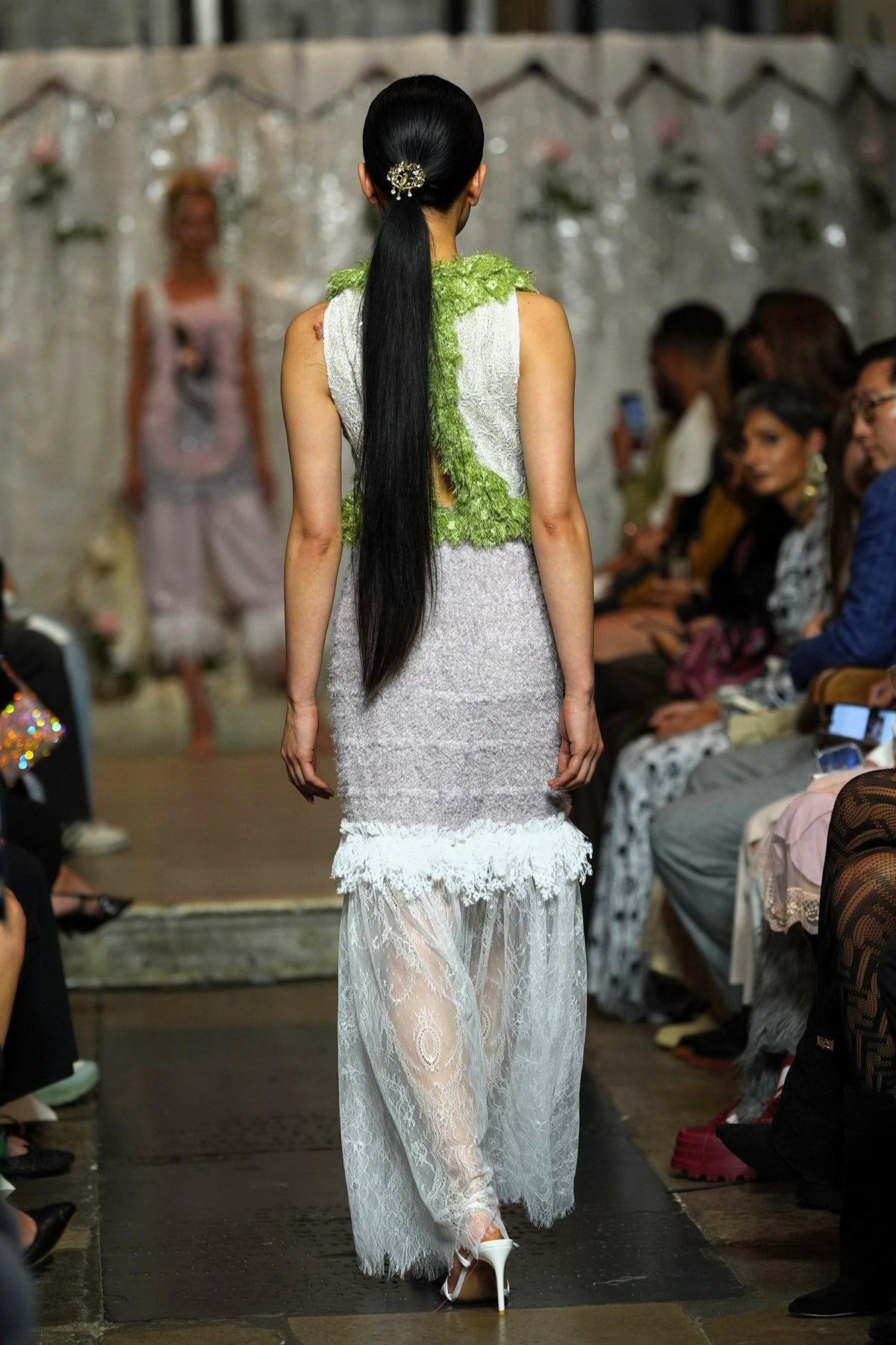 A model viewed from behind walking down a runway wearing a sleeveless white lace dress with a textured lime green bodice and a sheer white tulle overskirt. The runway is lined with spectators.