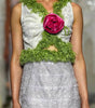 A model walks a fashion runway wearing a sleeveless white and light gray lace dress with a green floral collar and a large pink rose at the center chest.