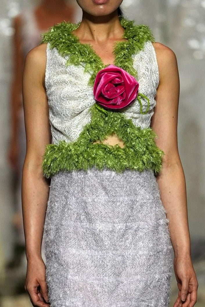 A model walks a fashion runway wearing a sleeveless white and light gray lace dress with a green floral collar and a large pink rose at the center chest.