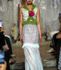 A model walks a fashion runway wearing a sleeveless white and light gray lace dress with a green floral collar and a large pink rose at the center chest.