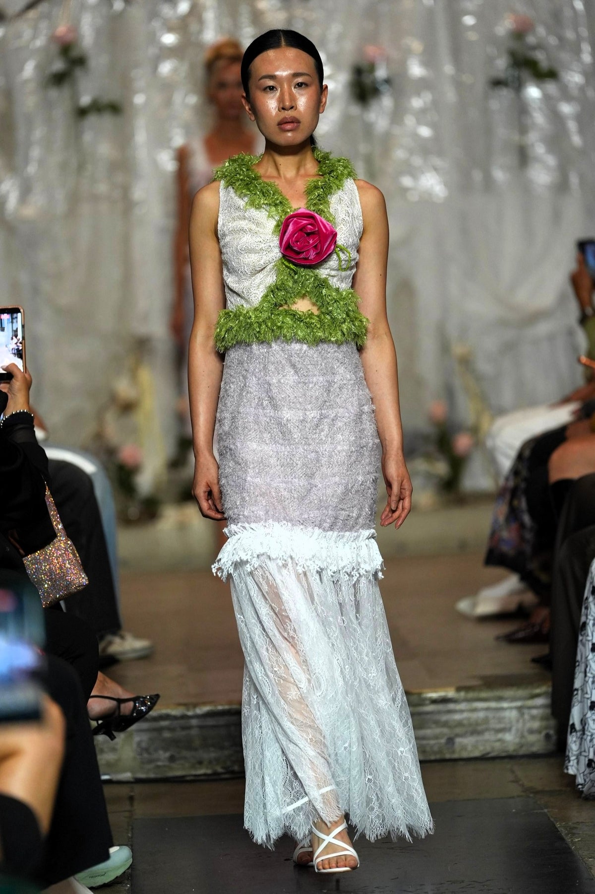 A model walks a fashion runway wearing a sleeveless white and light gray lace dress with a green floral collar and a large pink rose at the center chest.