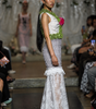 A model walks a fashion runway wearing a sleeveless white lace dress with a textured green floral waistband and a pink flower accent, set against a runway show background with seated spectators.