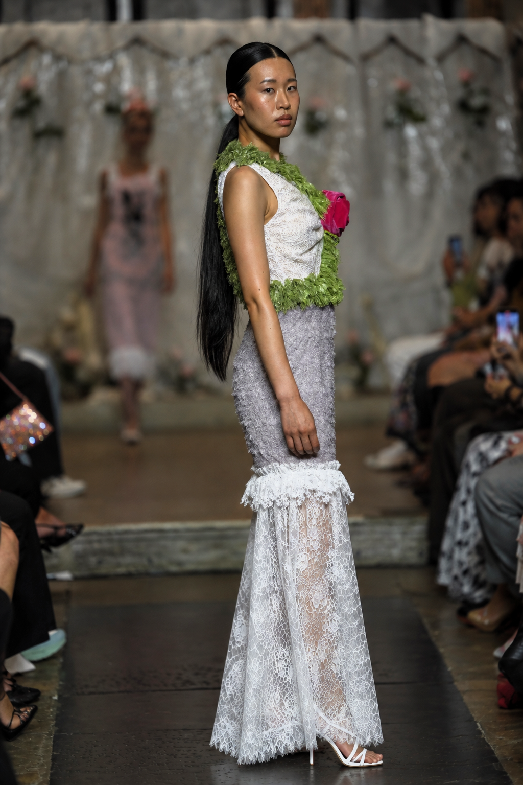 A model walks a fashion runway wearing a sleeveless white lace dress with a textured green floral waistband and a pink flower accent, set against a runway show background with seated spectators.