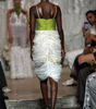 A model walks down a fashion runway wearing a sleeveless dress with a bright lime green bodice and a voluminous white ruffled skirt. The photo is taken from behind, with other runway attendees and a decorative backdrop visible in the background.