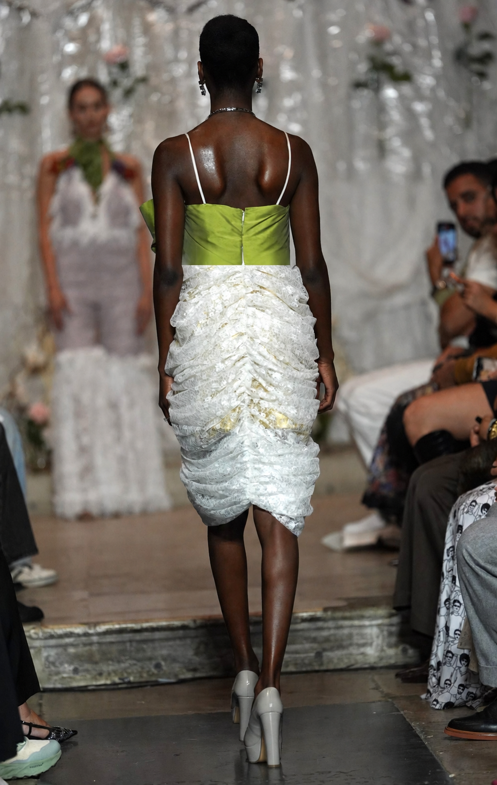 A model walks down a fashion runway wearing a sleeveless dress with a bright lime green bodice and a voluminous white ruffled skirt. The photo is taken from behind, with other runway attendees and a decorative backdrop visible in the background.