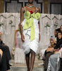 A runway model walks the catwalk wearing a white ruffled mini dress with a large lime green bow at the bust and a red rose accent, with an audience seated along the sides of the runway.