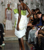 A model on a fashion runway wearing a white ruffled mini dress with a lime green bodice and white platform heels, with an audience seated along the runway.