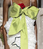 A runway model walks the catwalk wearing a white ruffled mini dress with a large lime green bow at the bust and a red rose accent, with an audience seated along the sides of the runway.
