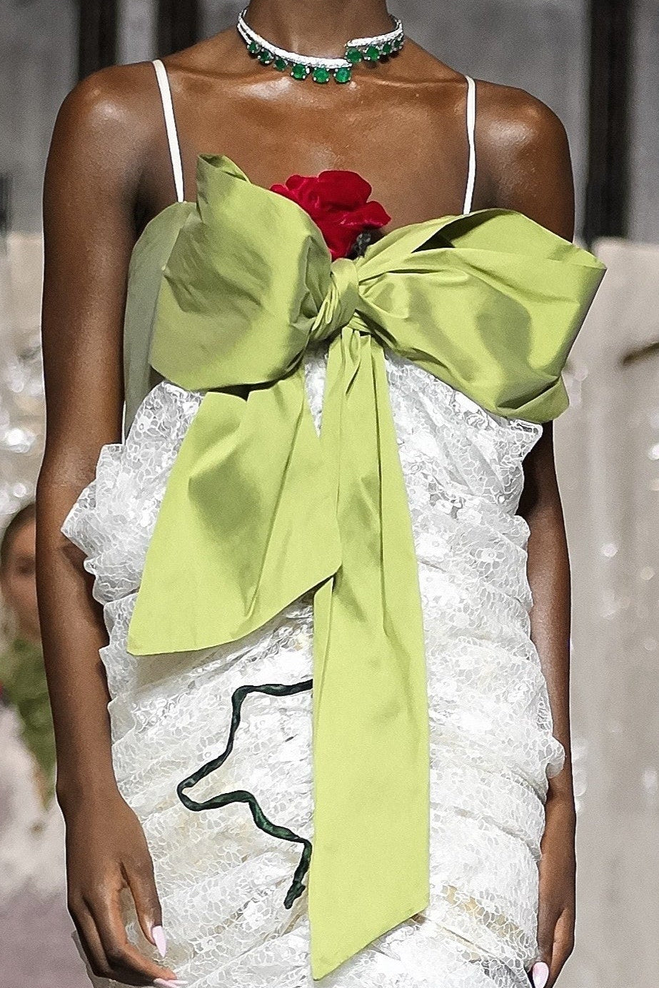 A runway model walks the catwalk wearing a white ruffled mini dress with a large lime green bow at the bust and a red rose accent, with an audience seated along the sides of the runway.