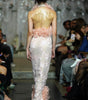 A model viewed from behind on a fashion runway wearing a sheer pale pink embellished gown with ruffled floral details at the waist and hem, with seated spectators along the catwalk.