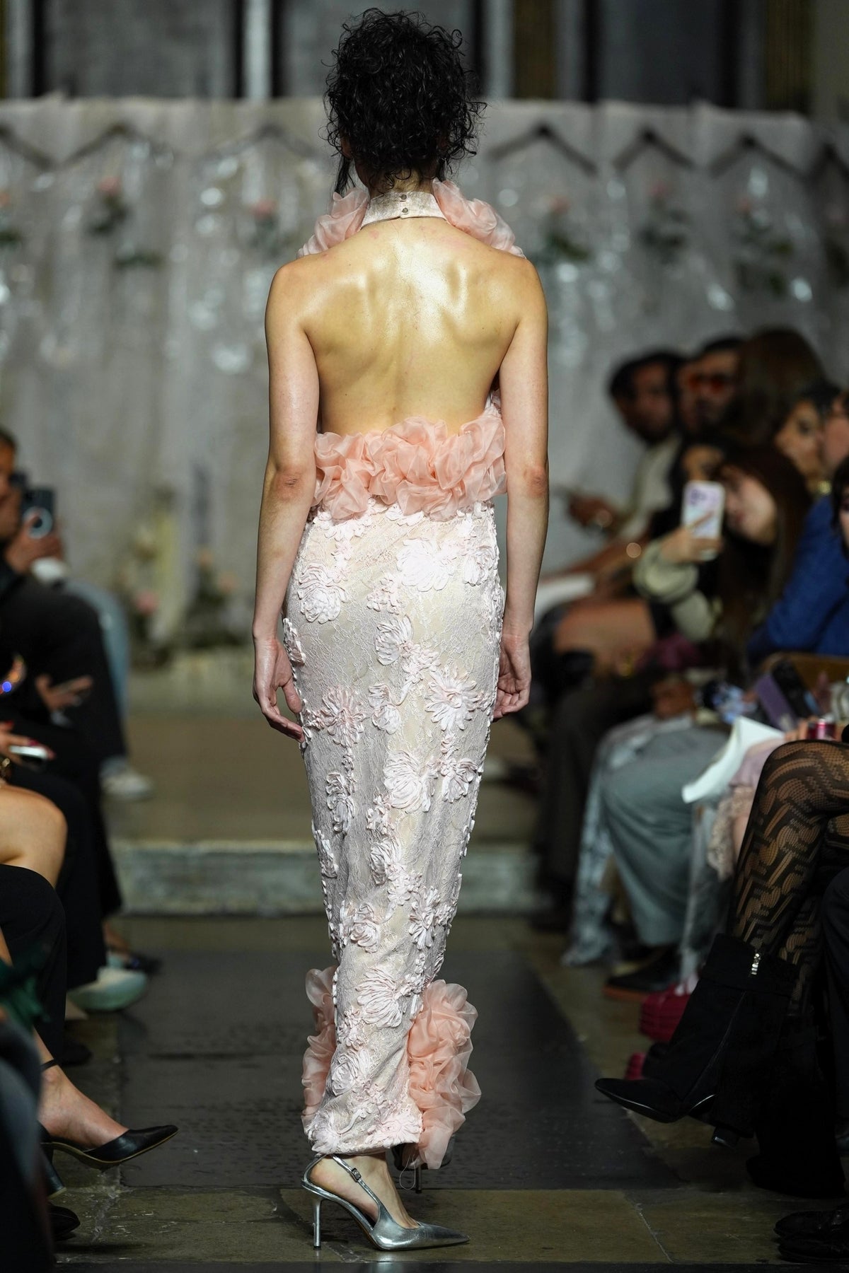 A model viewed from behind on a fashion runway wearing a sheer pale pink embellished gown with ruffled floral details at the waist and hem, with seated spectators along the catwalk.