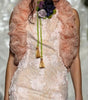 A model walks the runway wearing a pale blush pink floral lace maxi dress with a matching ruffled shrug and a floral chest accessory, set against a decorated white draped backdrop.