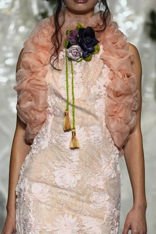 A model walks the runway wearing a pale blush pink floral lace maxi dress with a matching ruffled shrug and a floral chest accessory, set against a decorated white draped backdrop.
