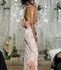 A model on a runway wearing a pale blush pink evening gown with a ruffled asymmetrical neckline, sheer floral lace skirt, and matching ruffled back detail, photographed during a fashion show.
