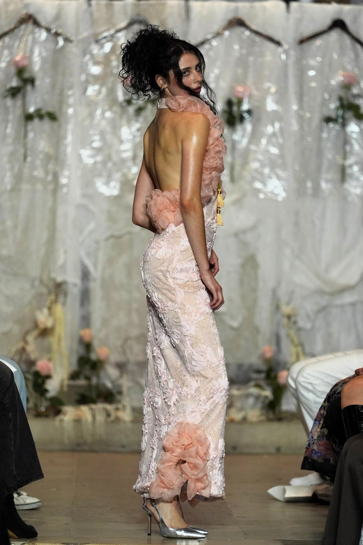 A model on a runway wearing a pale blush pink evening gown with a ruffled asymmetrical neckline, sheer floral lace skirt, and matching ruffled back detail, photographed during a fashion show.