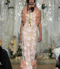 A model walks the runway wearing a pale blush pink floral lace maxi dress with a matching ruffled shrug and a floral chest accessory, set against a decorated white draped backdrop.