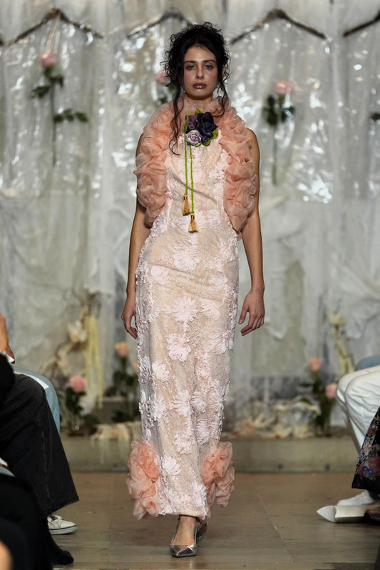 A model walks the runway wearing a pale blush pink floral lace maxi dress with a matching ruffled shrug and a floral chest accessory, set against a decorated white draped backdrop.