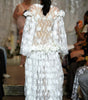 A model viewed from behind wearing a white, heavily textured, 3D floral lace gown on a fashion runway, with an audience seated in the background.