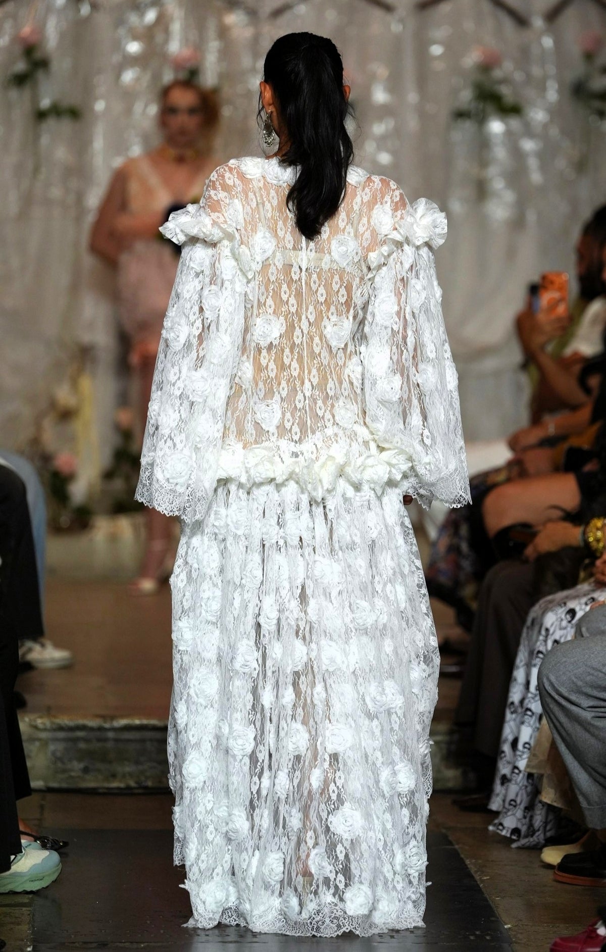 A model viewed from behind wearing a white, heavily textured, 3D floral lace gown on a fashion runway, with an audience seated in the background.