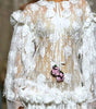 A model walks the runway wearing a long white sheer lace gown with 3D floral appliqué, dramatic ruffled sleeves, and a matching floral waist detail. Audience members are seated on both sides holding up phones to capture the show.