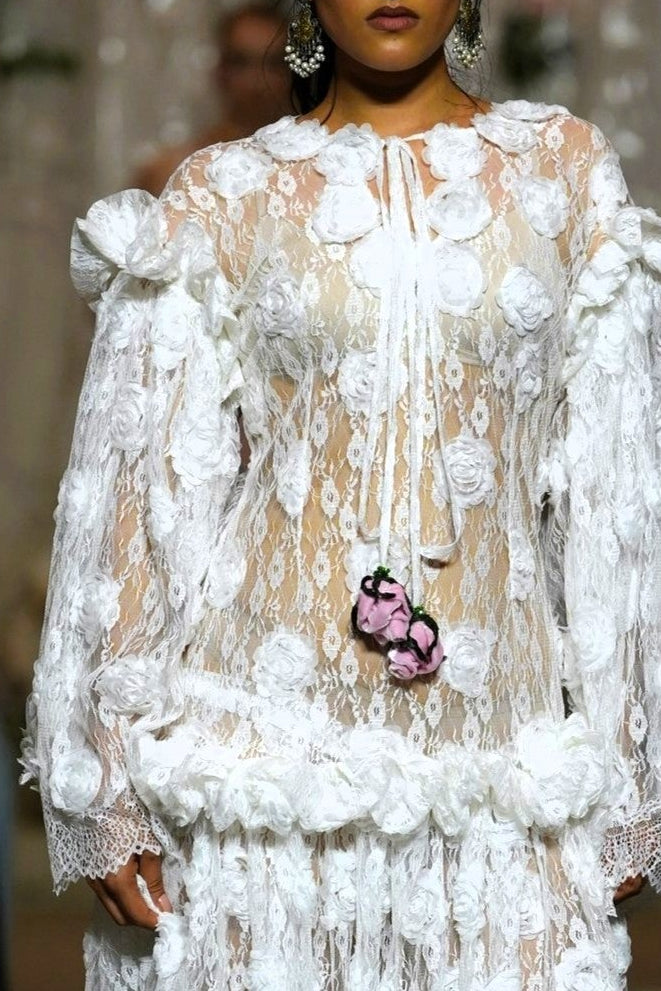 A model walks the runway wearing a long white sheer lace gown with 3D floral appliqué, dramatic ruffled sleeves, and a matching floral waist detail. Audience members are seated on both sides holding up phones to capture the show.