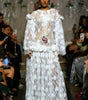 A model walks the runway wearing a long white sheer lace gown with 3D floral appliqué, dramatic ruffled sleeves, and a matching floral waist detail. Audience members are seated on both sides holding up phones to capture the show.