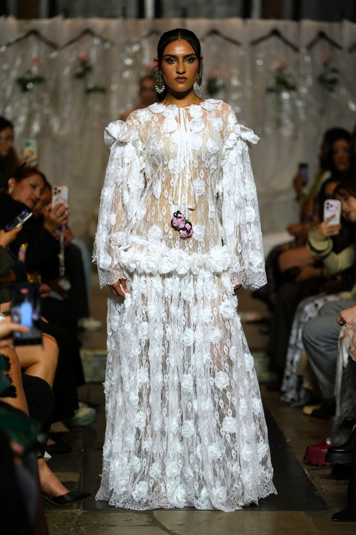A model walks the runway wearing a long white sheer lace gown with 3D floral appliqué, dramatic ruffled sleeves, and a matching floral waist detail. Audience members are seated on both sides holding up phones to capture the show.
