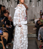 A model walks a fashion runway wearing a long white sheer lace dress with layered 3D floral appliqué and ruffled trim. The runway is surrounded by seated spectators holding up phones to capture the show.