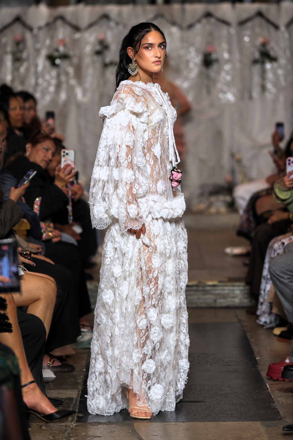 A model walks a fashion runway wearing a long white sheer lace dress with layered 3D floral appliqué and ruffled trim. The runway is surrounded by seated spectators holding up phones to capture the show.