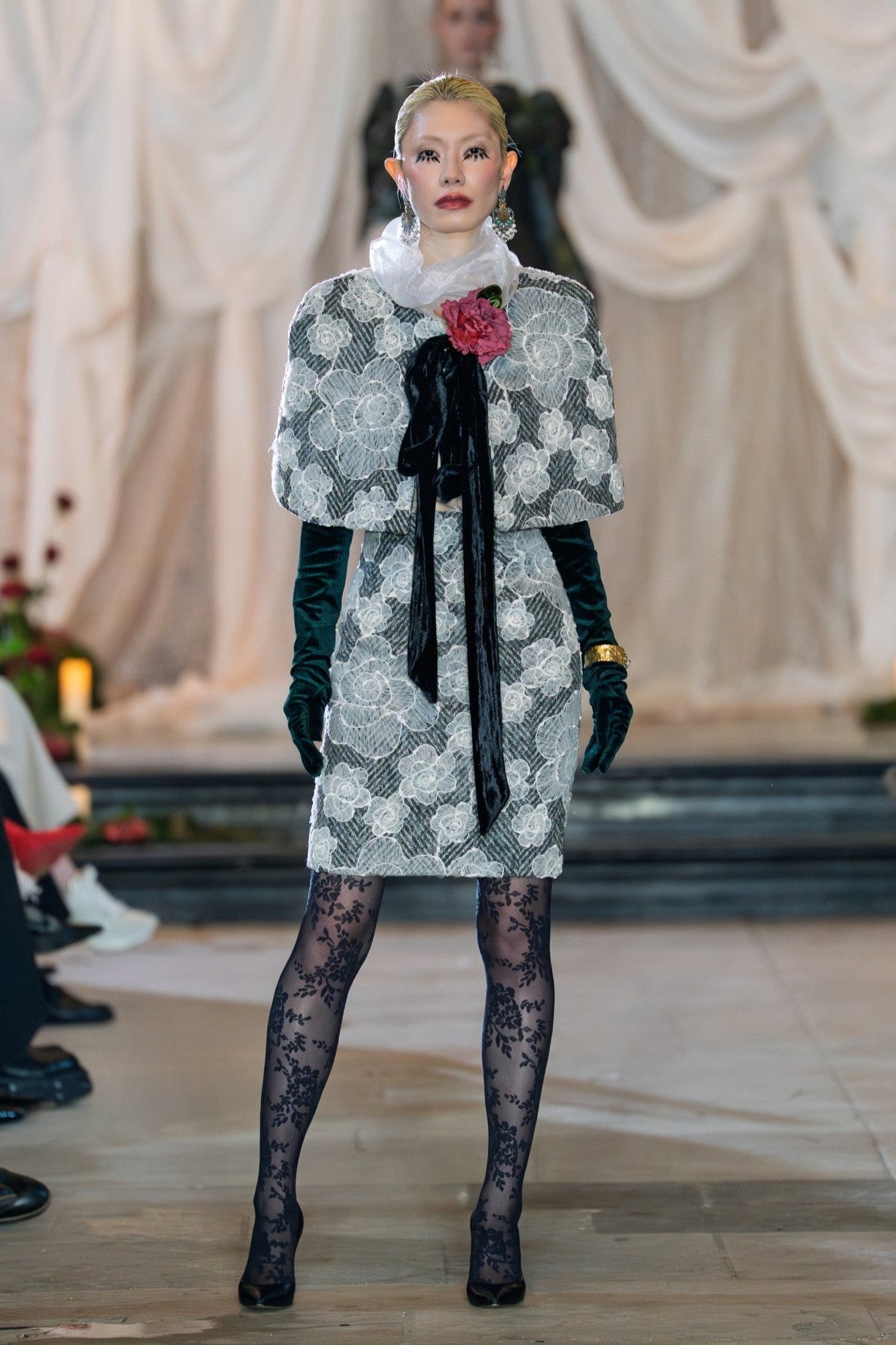 Model walking on a fashion runway wearing a patterned outfit with a floral accessory.