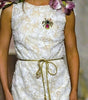 A model walks the runway wearing a sleeveless white lace midi dress with a gold tie belt, floral shoulder details, and a brooch at the neckline.