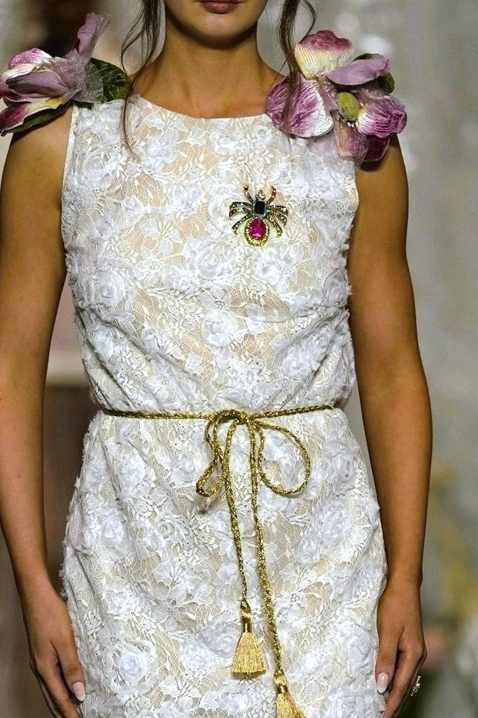 A model walks the runway wearing a sleeveless white lace midi dress with a gold tie belt, floral shoulder details, and a brooch at the neckline.