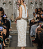 A model on a runway wearing a sleeveless, textured cream lace midi dress with a draped shoulder detail and nude high heels, with seated spectators on both sides.